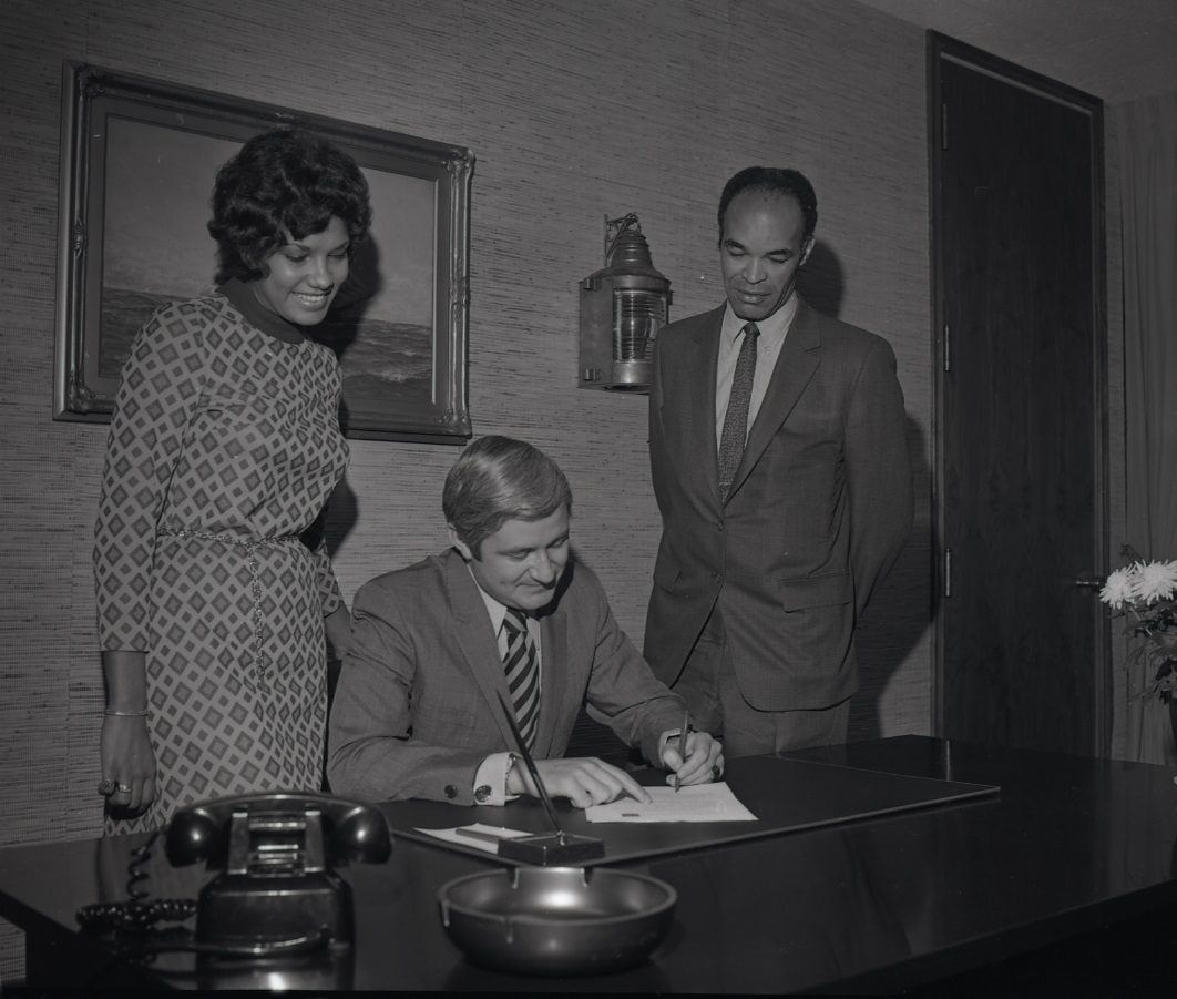 Black and white picture from 1971 of two people standing behind a person seated at a desk signing a document