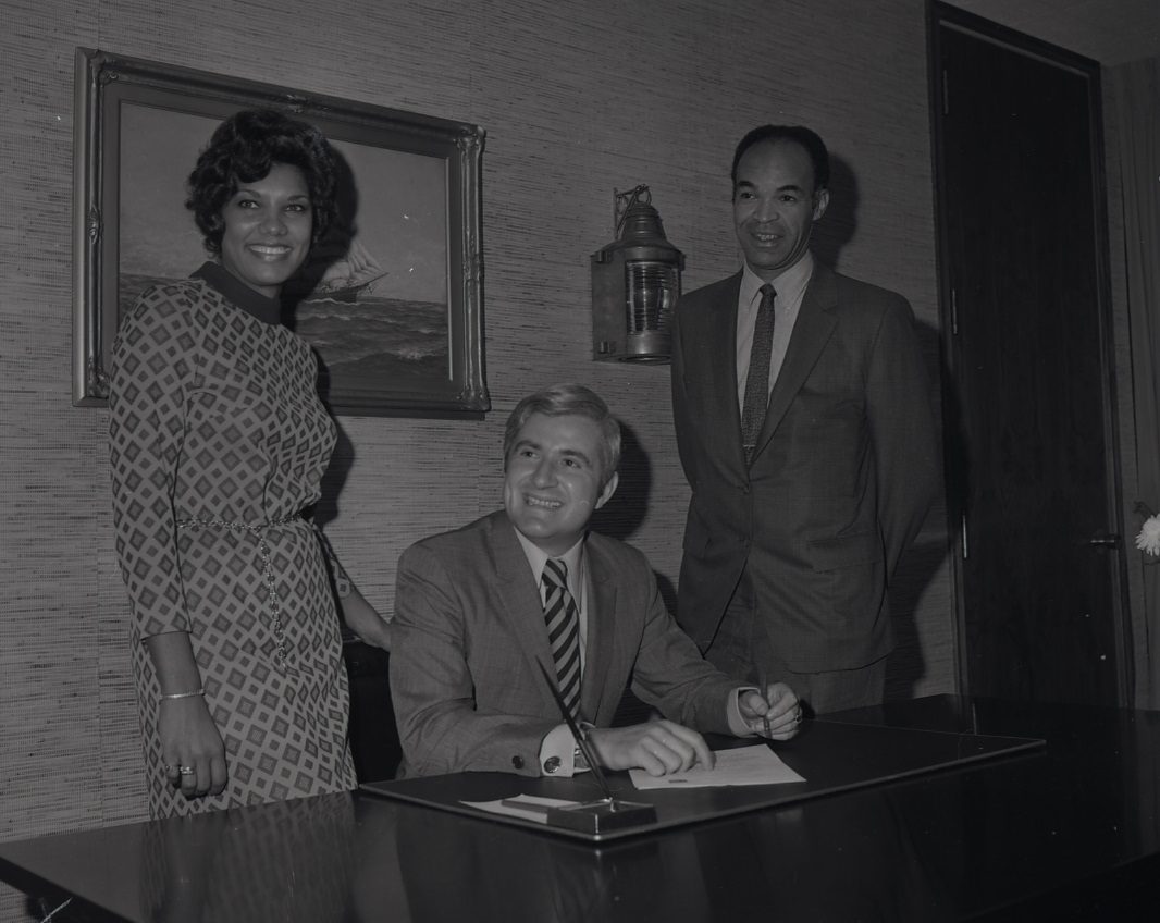 Black and white picture from 1971 of two people standing behind a person seated at a desk signing a document