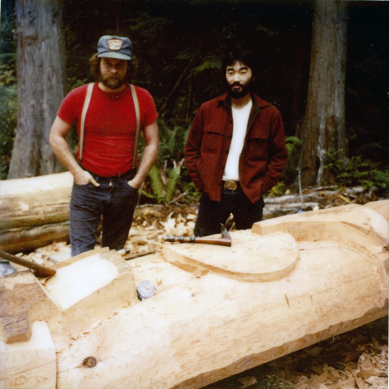 color photo of two men standing next to a totem pole in the process of being carved