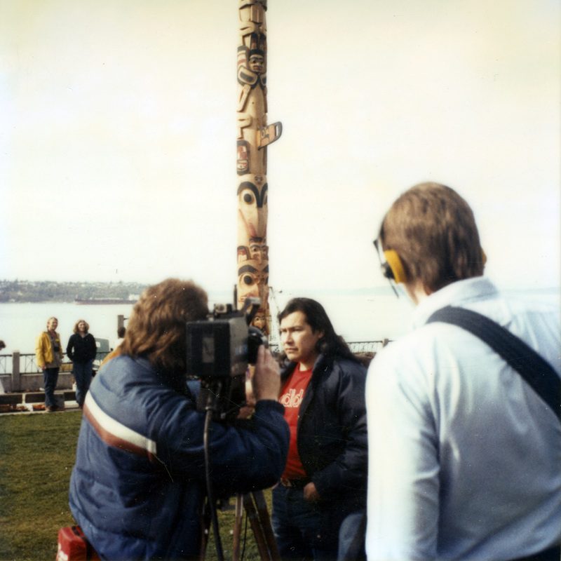color photo of totem pole carver Marvin Oliver talking to the press in Market Park in front of a totem pole