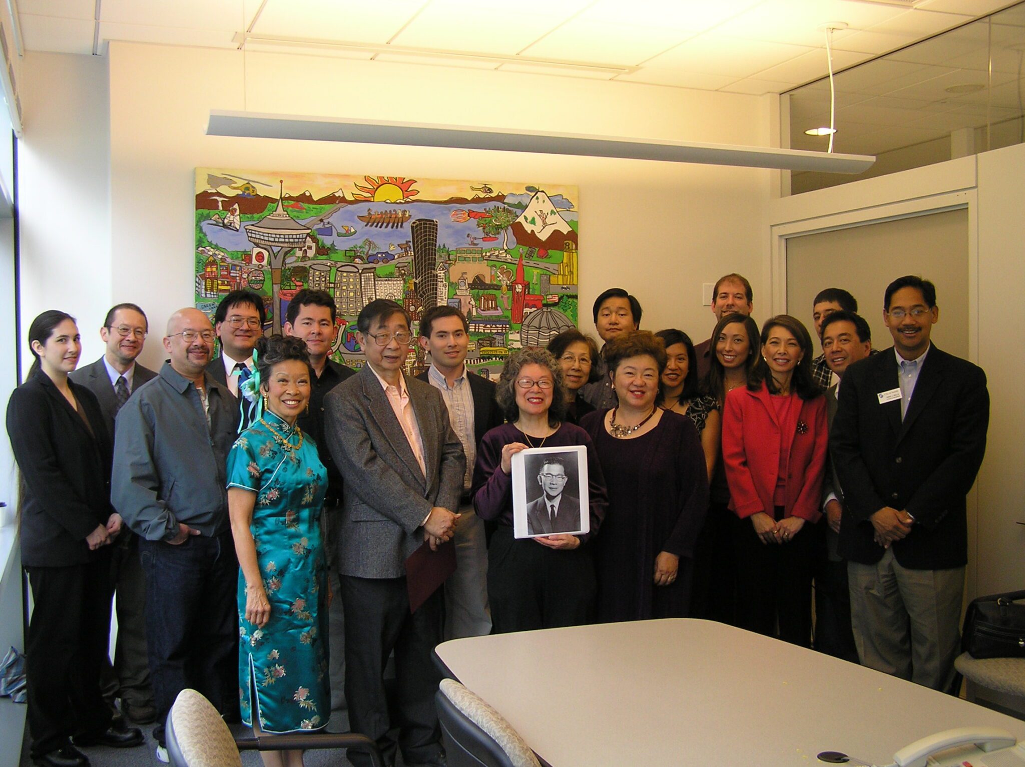 Color photo showing a group of people in the Wing Luke conference room at Seattle City Hall standing around a photo of former Councilmember Wing Luke.