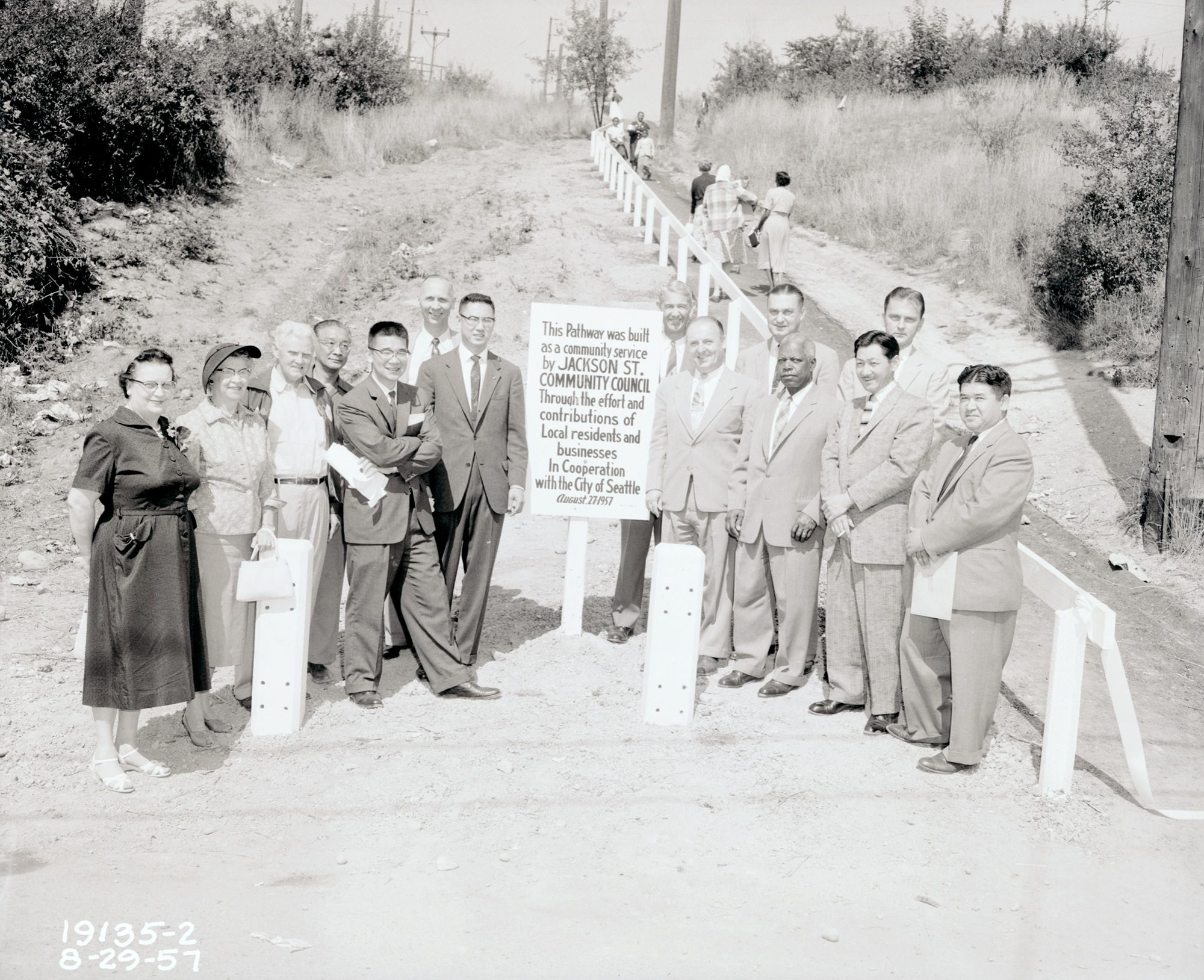 Black and white photo of a group of people at the base of a path leading up a hill, surrounding a sign reading: This Pathway was built as a community service by Jackson St. Community Council Through the effort and contributions of Local residents and businesses in Cooperation with the City of Seattle 