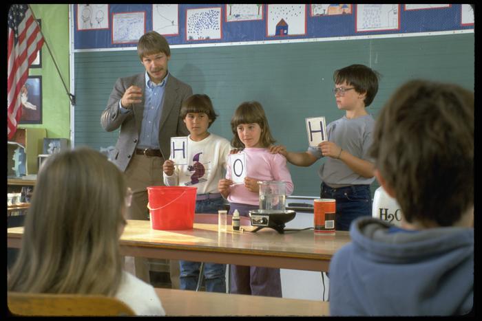 Man with three children standing at front of classroom. Two children are holding "H" signs and one has an "O."