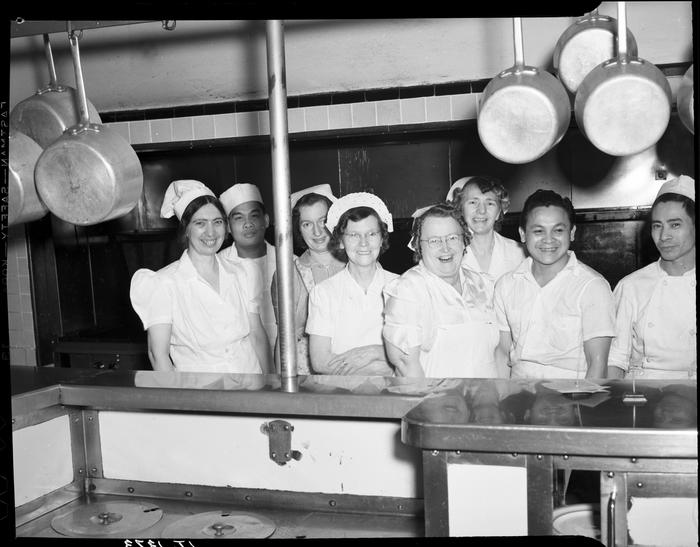 Eight people in uniforms posing behind commercial kitchen counter with pans hanging above