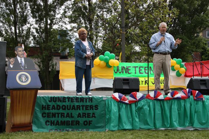 Mayor Mike McGinn on a stage with an unidentified woman; a Juneteenth banner and balloons are in the background and a cardboard cutout of President Obama speaking is at left