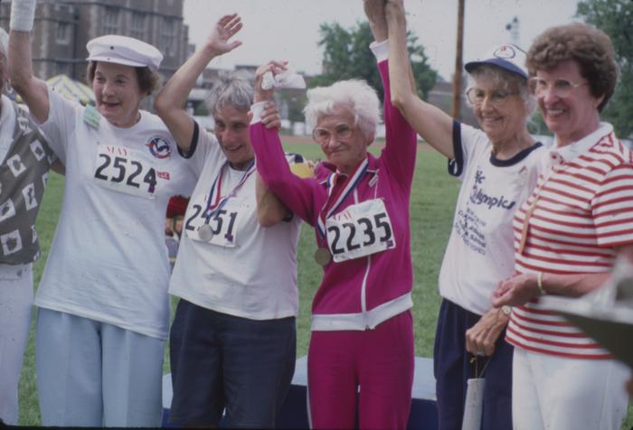 Five senior women raising arms above their heads. Three are wearing race numbers and two have medals.