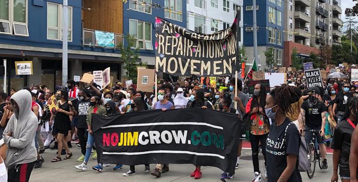 Crowd marching in street with a banner reading "NoJimCrow.com" and another only partly legible with text including "repair-ations" and "movement"
