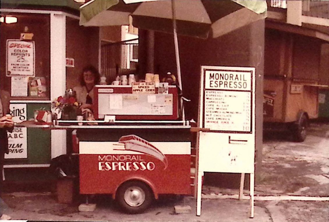 Woman standing behind small sidewalk cart labeled "Monorail Espresso." A sign that appears to be a menu board stands next to the cart.
