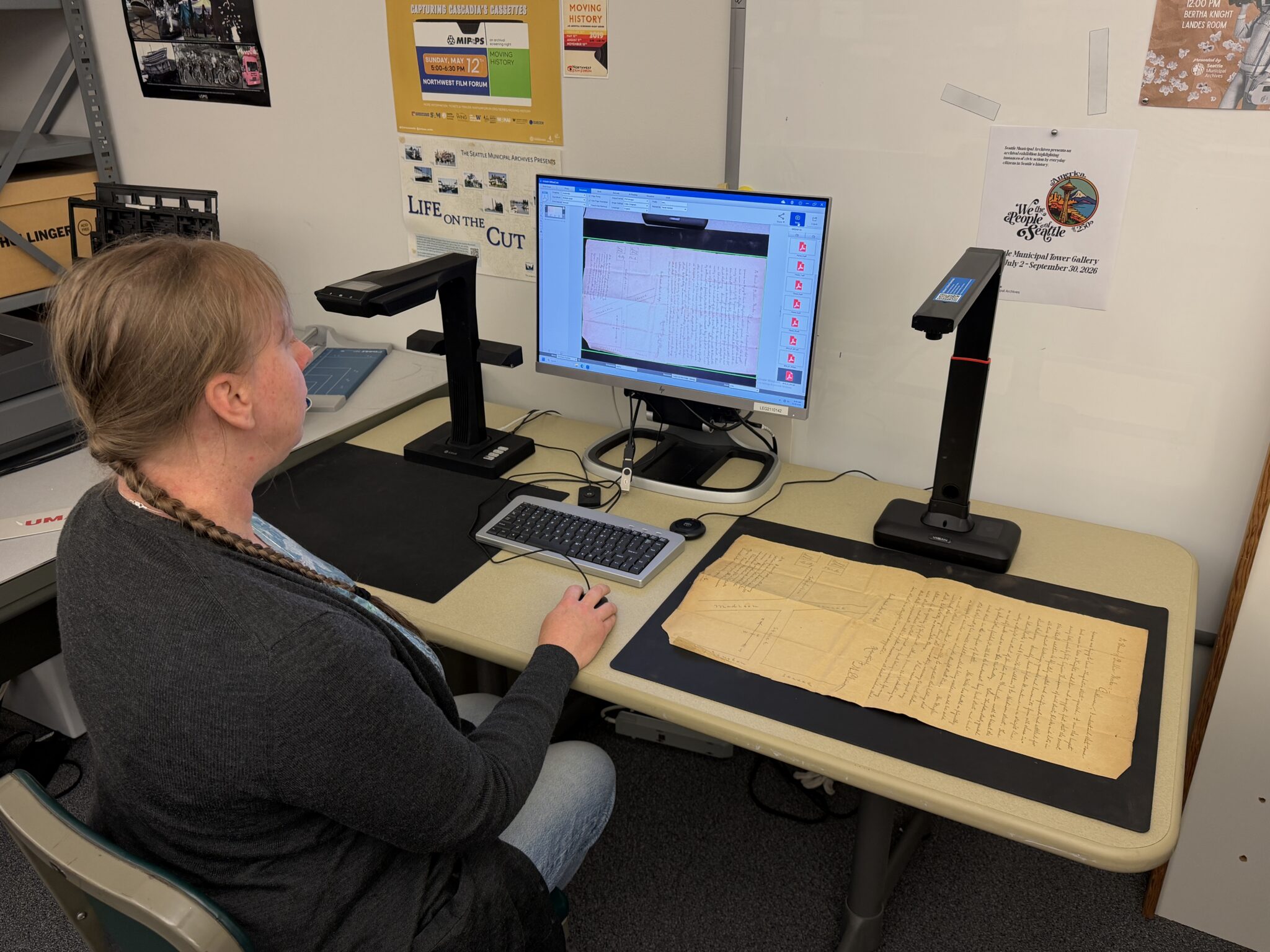 woman seated at computer with a 19th century document flat under an overhead scanner